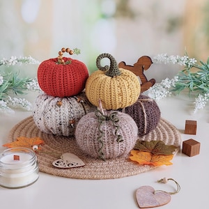 Puede incluir: Una colección de cinco calabazas de ganchillo en rojo, amarillo, crema con motas negras, morado claro y marrón oscuro. Cada calabaza presenta un tallo único hecho de cuentas, hilo o cordel. Las calabazas están dispuestas sobre un mantel de yute.