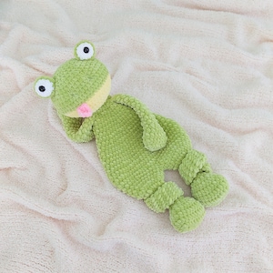 Könnte beinhalten: Ein handgefertigtes grünes Häkelfrosch-Plüschtier mit großen, runden Augen und einer rosa Zunge. Der Frosch liegt auf einer weichen, hellrosa Decke. Das Plüschtier hat ein strukturiertes, flauschiges Aussehen.