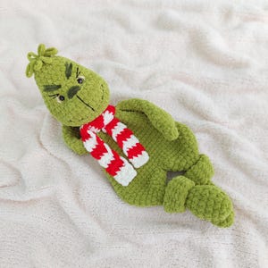 Puede incluir: Muñeco Grinch verde tejido a ganchillo con una bufanda a rayas rojas y blancas. El muñeco tiene una expresión gruñona, con detalles negros cosidos para los ojos, las cejas y la boca. El muñeco está sobre una superficie blanca texturizada.