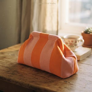 May include: A soft, knitted pouch featuring vertical stripes in alternating shades of bright orange and light pink. The bag has a structured, kiss-lock style frame closure at the top and a textured, fabric-like appearance throughout its exterior.