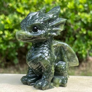 May include: A carved dark green stone figurine of a small dragon with large eyes, pointed ears, and textured scales. The creature is depicted in a seated position with folded wings, showcasing a polished surface and deep forest green colour tones.