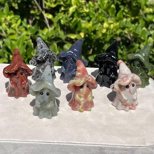 Puede incluir: Una colección de ocho pequeñas figuras de piedra talladas a mano con forma de fantasmas que llevan sombreros puntiagudos. Cada fantasma presenta un acabado liso y pulido, además de dos pequeños ojos circulares oscuros. Las figuras están hechas de diversas piedras en colores rojo, gris, azul, negro, verde y blanco con vetas rosas.