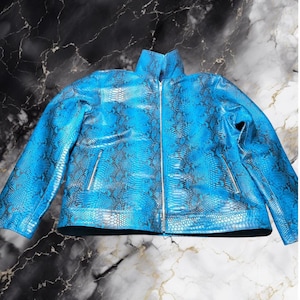Könnte beinhalten: Eine leuchtend blaue Jacke mit einem durchgehenden schwarzen Schlangenhautmuster. Die Jacke hat einen hohen Kragen, lange Ärmel und einen durchgehenden silberfarbenen Reißverschluss. Auf der Vorderseite befinden sich zwei vertikale Reißverschlusstaschen.