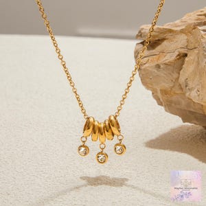 May include: A gold-toned necklace featuring a delicate chain with five smooth, rounded gold-toned beads suspended from the center. Three of these beads are accented with small, clear, round faceted crystals hanging from the bottom of the chain.