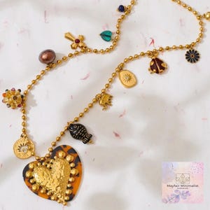 Può includere: Collana dorata con vari ciondoli, tra cui un cuore, un pesce, una croce e una coccinella. Il ciondolo a forma di cuore ha un centro dorato strutturato e un bordo con motivo tartarugato. La collana è su una superficie bianca.