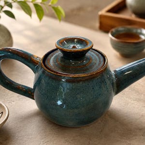 May include: A teal ceramic teapot with a matching lid and handle. The teapot has a brown rim and spout. A small bowl of tea leaves and two teacups are also visible. The teapot is part of a tea set.