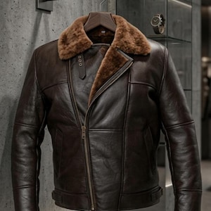 May include: A dark brown leather jacket featuring a thick, plush tan shearling collar and interior lining. The jacket includes an asymmetrical front zipper closure, long sleeves, and a small buckled strap detail near the collar neckline.