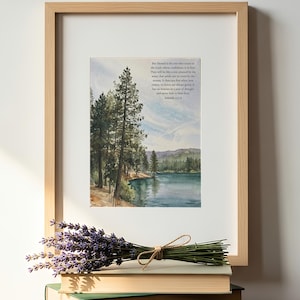 May include: A framed watercolor print featuring a serene lake surrounded by tall pine trees and mountains. The text of Jeremiah 17:7-8 is printed in the upper right corner. A bundle of dried purple lavender rests on a stack of three books.