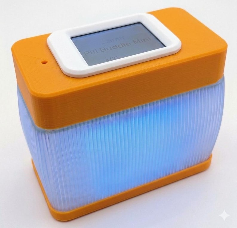Puede incluir: Un Pill Buddie Mini naranja y blanco con una peque&ntilde;a pantalla que muestra texto. El dispositivo tiene forma rectangular con una luz azul brillante que emana de la mitad inferior. El texto "Pill Buddie Mini" es visible en la pantalla.