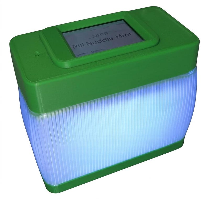 Puede incluir: Un dispositivo rectangular verde con una peque&ntilde;a pantalla que muestra "Pill Buddie Mini". La parte inferior del dispositivo es transl&uacute;cida y emite un brillo azul. El dispositivo tiene una forma redondeada.