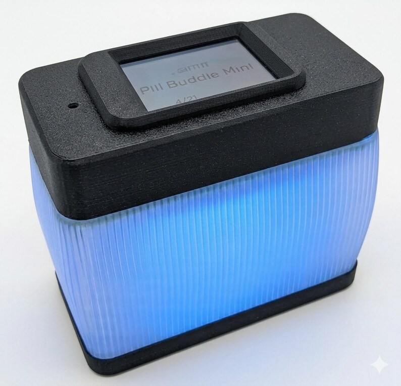 Puede incluir: Un dispositivo rectangular negro con una peque&ntilde;a pantalla que muestra "Pill Buddie Mint". La parte inferior del dispositivo brilla con una luz azul brillante. El dispositivo tiene un cuerpo transl&uacute;cido y acanalado.