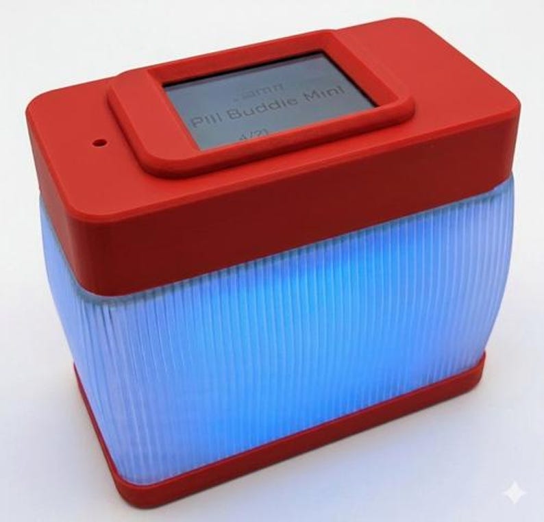 Puede incluir: Un dispositivo rectangular rojo con una peque&ntilde;a pantalla que muestra "Pill Buddie Mini". La mitad inferior es transl&uacute;cida con l&iacute;neas verticales, iluminada por una luz azul. Dise&ntilde;o moderno.