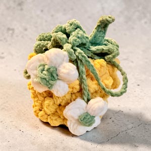 May include: A small, round, yellow crocheted pouch featuring a drawstring closure with green yarn ties. The exterior is decorated with two white crocheted flowers that have green centers, adding a textured, layered appearance to the design.