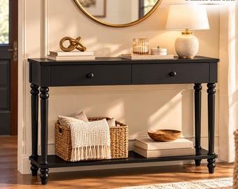 May include: A black console table with two drawers and four turned legs, featuring a lower shelf holding a woven basket with a white knit throw and a stack of books topped with a wooden bowl. The top surface holds a lamp and decor.