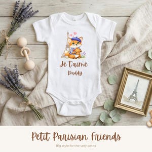 May include: White baby onesie with a watercolor illustration of a fox in a beret in front of the Eiffel Tower. The text "Je t'aime Daddy" is printed below the fox. The words "Petit Parisian Friends" are at the bottom.