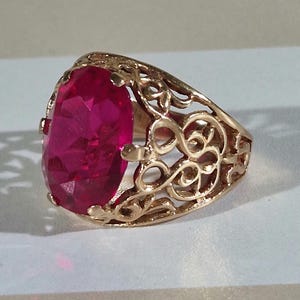 Puede incluir: Un anillo de tono dorado con una gran piedra preciosa de color rosa vibrante y corte ovalado, engastada en una banda de metal decorativa y calada con intrincados patrones de remolinos. La piedra facetada está centrada y es muy brillante.