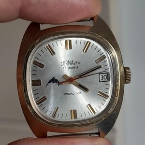 May include: Vintage Cornavin watch with a silver face, gold-tone case, and black hands. The watch features a dolphin logo, the number 30, and the text "shockproof". The watch is held by a person's fingers.