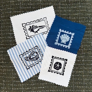 May include: Four rectangular fabric patches with pinked edges featuring stamp-style prints. One white patch shows a black conch shell, one blue patch shows a white scallop shell, one white patch shows a black sand dollar, and one blue-striped patch shows a black oyster.