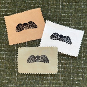 May include: Three rectangular fabric swatches with pinked edges, each featuring a black ink print of a winged skull. The swatches are in tan, white, and light olive green, displaying a consistent graphic design across all three pieces of fabric.