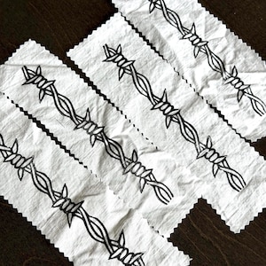 May include: Three rectangular white fabric strips with pinked edges, each featuring a continuous black ink illustration of barbed wire running diagonally across the center. The design shows twisted metal strands with sharp, pointed barbs.