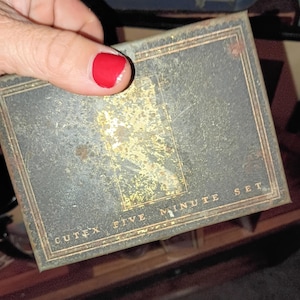 May include: A vintage Cutex Five Minute Set box. The box is rectangular with a dark grey and gold colour scheme. The text "CUTEX FIVE MINUTE SET" is printed on the front. The box is held by a hand with red nail polish.