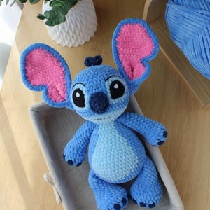 May include: A handmade blue crochet plush toy with large pink-lined ears, black eyes, and a dark blue nose. The plush is in a gray basket on a wooden surface. The toy is a character from a popular animated film.