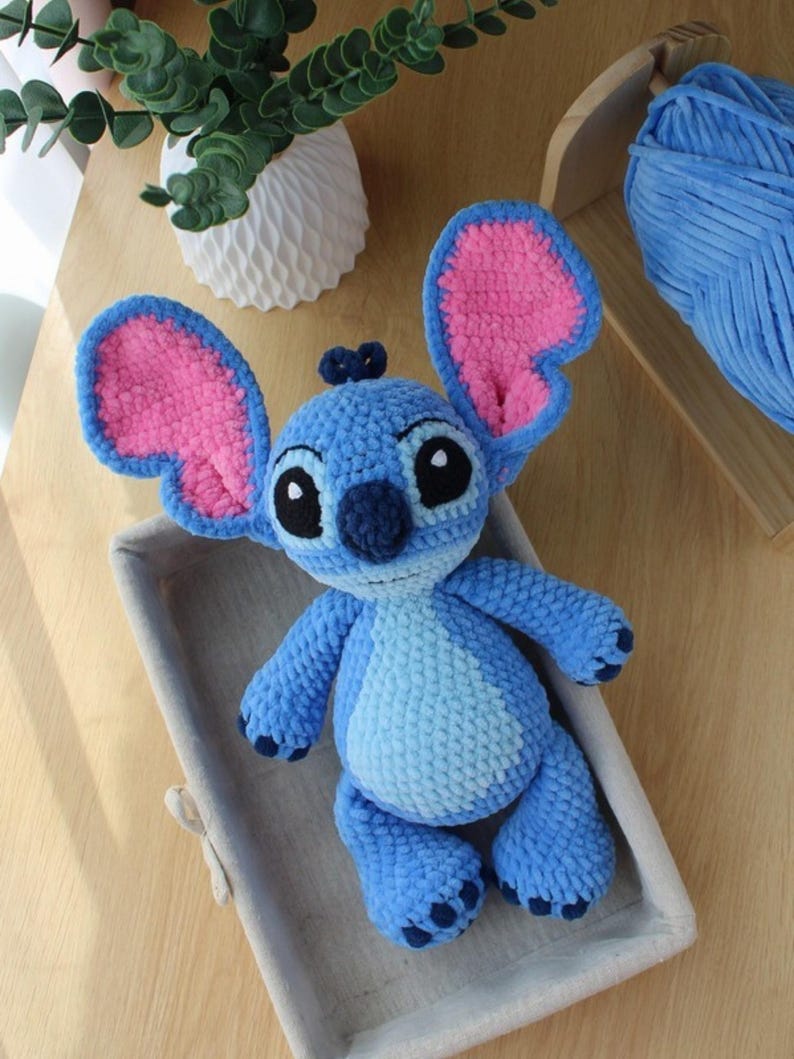 May include: A handmade blue crochet plush toy with large pink-lined ears, black eyes, and a dark blue nose. The plush is in a gray basket on a wooden surface. The toy is a character from a popular animated film.