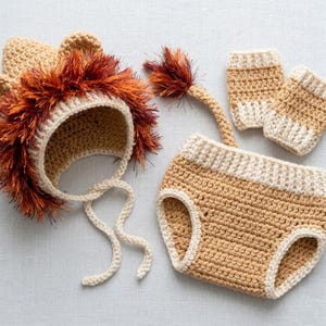 May include: A crochet lion costume set consisting of a tan bonnet with a textured rust-colored fringe mane and ear details, a pair of tan and cream wrist cuffs, and a tan diaper cover featuring an attached tail with a rust-colored tuft.