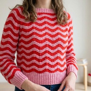 May include: A long-sleeved knit jumper featuring a horizontal wavy pattern in alternating bright red and light pink stripes. The garment has a round neckline and ribbed cuffs and hem in a solid light pink colour, constructed with a textured knit.