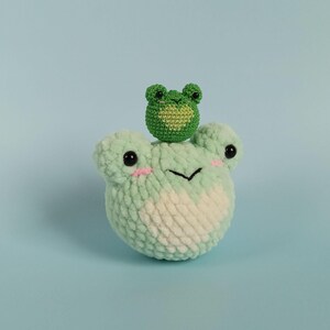 May include: A handmade crochet frog with a smaller frog on its head. The larger frog is light green with a white belly, black eyes, and pink cheeks. The smaller frog is dark green. The background is light blue.