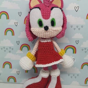May include: A handmade crochet doll of a character with pink fur, green eyes, and a red dress. The doll has white gloves, yellow bracelets, and red boots with white accents. The background features rainbows and hearts.