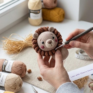 Op de afbeelding: Een gehaakt leeuwtje met een beige gezicht, donkerbruine manen en roze wangen. De leeuw wordt bewerkt met een grijze haaknaald. In de buurt liggen bollen garen in beige, bruin en geel, een metalen schaar en een papieren label.