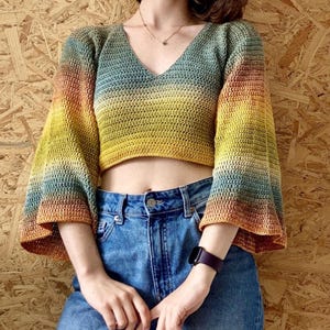 Puede incluir: Un top de ganchillo corto con cuello en V y mangas acampanadas. El top presenta un degradado de colores, que va del naranja al amarillo, verde y azul. El top se combina con unos vaqueros azules.