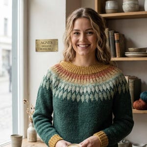 May include: A long-sleeved knit jumper featuring a dark green base with a circular yoke pattern in shades of mustard yellow, rust orange, and light green. A brass plaque on the wall displays the text AGNES SWEATER MODEL NO. 1 in black lettering.