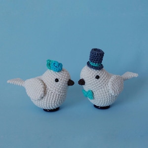 May include: Two white crochet birds with black eyes and brown beaks. One bird wears a blue top hat and bow tie, the other a blue flower. The birds are on a blue background, ideal for wedding decor.