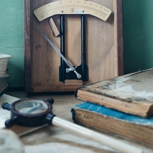 Può includere: Un dispositivo di misurazione a parete in legno vintage con telaio verticale in metallo nero, una scala beige curva graduata da 0 a 100 e un sottile puntatore in metallo. La scala riporta il testo 1965 e OCT 40115. Una piccola lente d'ingrandimento è vicina.