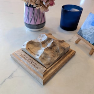 Puede incluir: Escultura de montaña de madera con una base marrón claro y una cordillera tallada en detalle. La base tiene el texto "Mountain Height Personalised Message". Un jarrón con flores rosas y un vaso azul en el fondo.