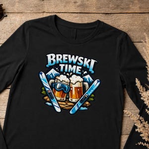 May include: A black long-sleeve t-shirt featuring a graphic print of two beer mugs, blue ski goggles, and two skis. The text BREWSKI TIME is printed in white and light blue above the mugs, with snowy mountain peaks in the background.