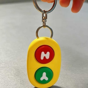 Puede incluir: Un llavero ovalado de color amarillo con dos botones circulares. El botón superior es rojo con una letra H blanca y el botón inferior es verde con una letra Y blanca. El llavero está unido a una anilla de metal plateado mediante un cierre.