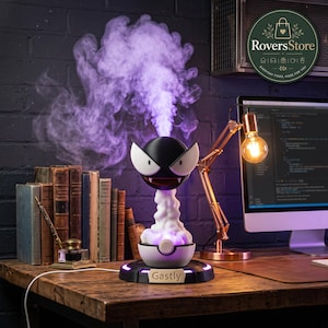 May include: A black and white Gastly-themed humidifier emitting purple vapor, resting on a circular base with the text Gastly. The device features a spherical head with large white eyes and a fanged mouth, positioned above a white and black base.