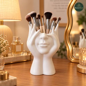 Peut inclure: Un organisateur de bureau en céramique blanche en forme de mains entourant un visage, rempli de divers pinceaux de maquillage. La sculpture présente une finition mate lisse et représente un visage humain aux yeux et lèvres fermés.