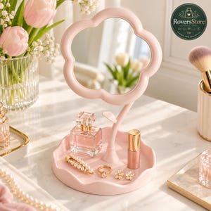 May include: A light pink vanity mirror shaped like a flower with a matching circular tray base. The mirror stand features a small leaf detail. The tray holds a square glass perfume bottle, a pearl hair clip, a gold ring, and two flower earrings.