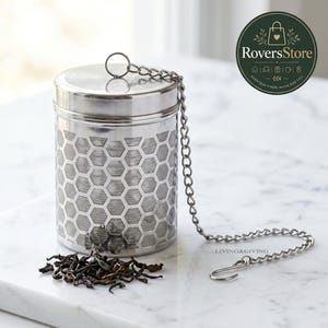 May include: A cylindrical stainless steel tea infuser featuring a laser-cut honeycomb pattern on its body, a secure lid, and a long metal chain with a hook at the end. A small pile of loose-leaf black tea sits next to the infuser on the surface.