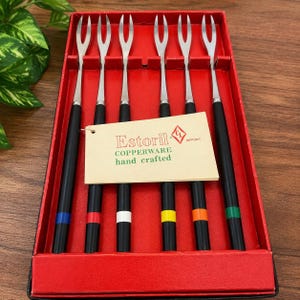 May include: A set of six two-pronged metal forks with black handles, each featuring a distinct colored ring in blue, red, white, yellow, orange, or green. The forks are arranged in a red box with a tag labeled Estoril Copperware hand crafted.