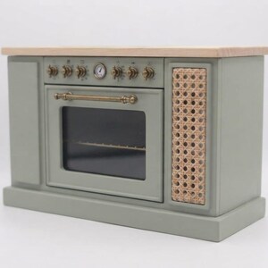 May include: A sage green toy oven featuring a light wood countertop, a central glass-paneled door with a brass-toned handle, and a side panel with a natural woven cane texture. The control panel includes brass-toned knobs and a small circular gauge.