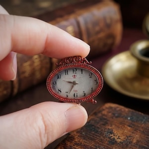 May include: A small, antique-style clock with a red oval frame. The clock face is white with black numerals and hands. The word "Paris" is printed at the bottom. The clock is held between fingers.