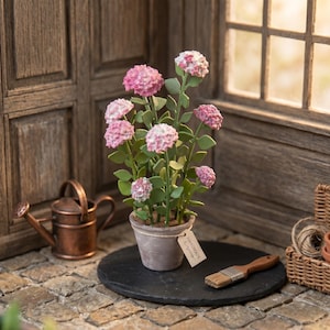Op de afbeelding: Een miniatuur hortensia in pot met roze en witte bloemen. De plant staat op een zwart stenen oppervlak, naast een kleine koperen gieter en een penseel. Een label op de pot leest "Handmade".