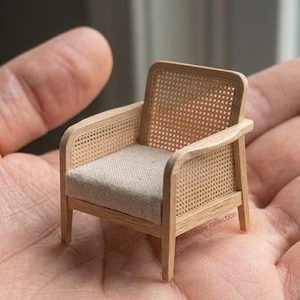 Könnte beinhalten: Ein Miniatursessel mit einem hellen Holzrahmen und einer Rückenlehne sowie Seitenteilen aus geflochtenem Rohrgeflecht. Die Sitzfläche ist mit einem schlichten, hellbeigen Stoff bezogen. Der Stuhl ist klein genug, um in einer Handfläche zu liegen.