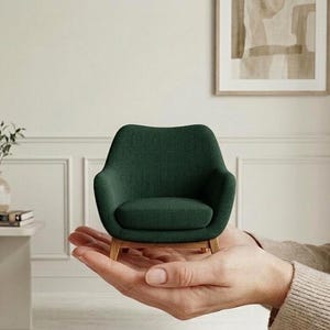 Könnte beinhalten: Ein Miniatur-Sessel mit dunkelgrünem Stoffbezug, geschwungener Rückenlehne, abgerundeten Armlehnen und hellen Holzbeinen. Das kleine Möbelstück wird in der Handfläche gehalten und zeigt seine kompakte Größe sowie das detaillierte Design.