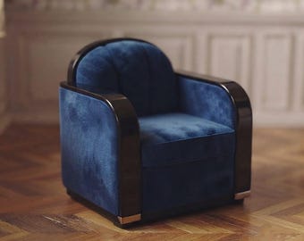 Könnte beinhalten: Ein Miniatursessel mit einem Bezug aus tiefblauem Samtstoff. Der Sessel verfügt über eine abgerundete Rückenlehne und dunkel polierte Holzarmlehnen, die nach unten gebogen sind und an der Basis mit kleinen Metalldetails verziert sind.